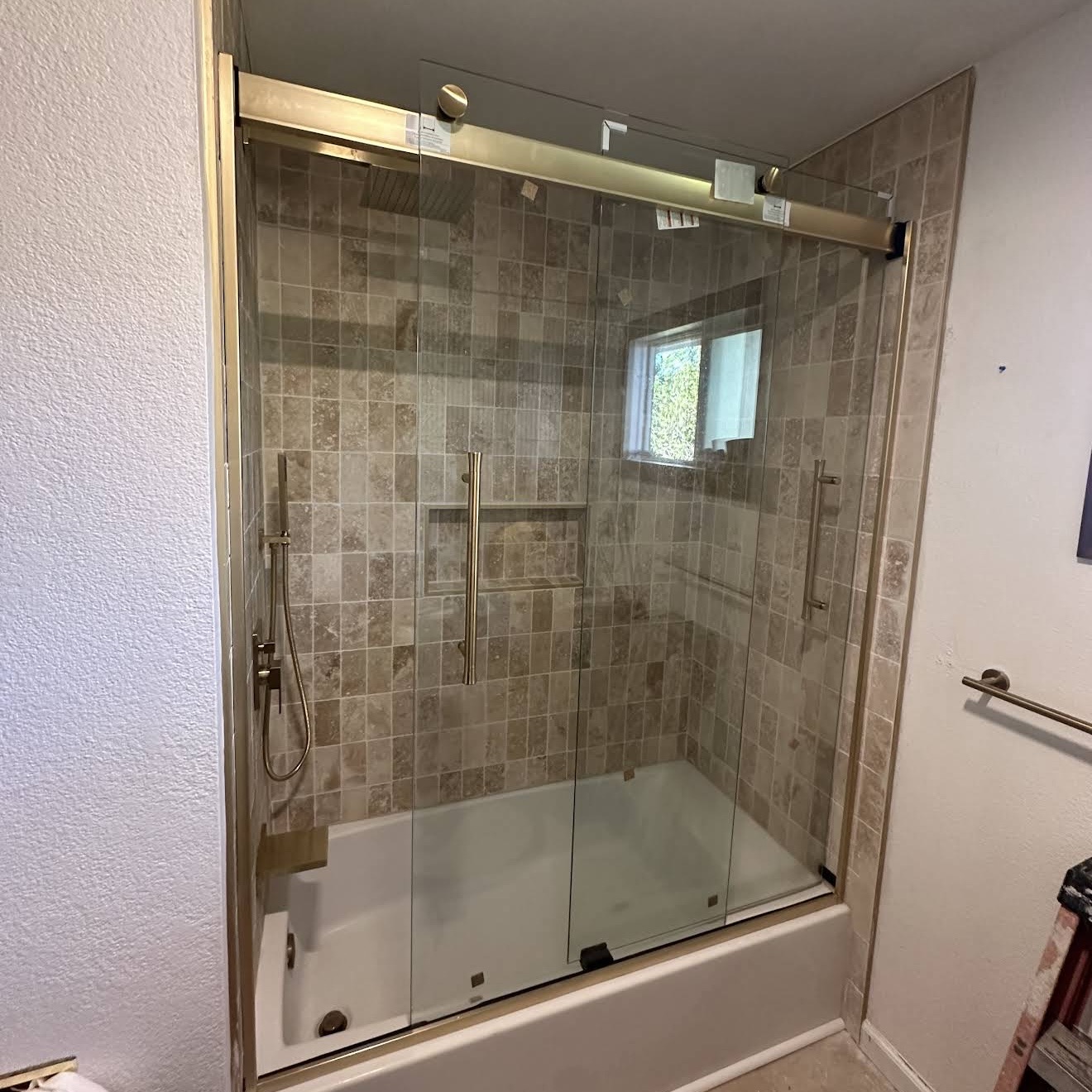 Frameless glass shower with brushed brass fixtures over tan stone tile.